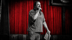 ChadO performing stand up comedy at Tank’s Pizza in San Antonio while holding handwritten notes on a yellow legal pad.